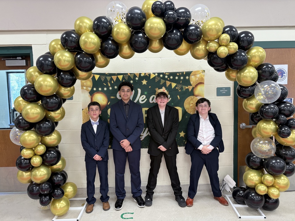 Four people stand together under a large balloon arch. They wear formal suits and shoes. The arch is black, gold, and white.
