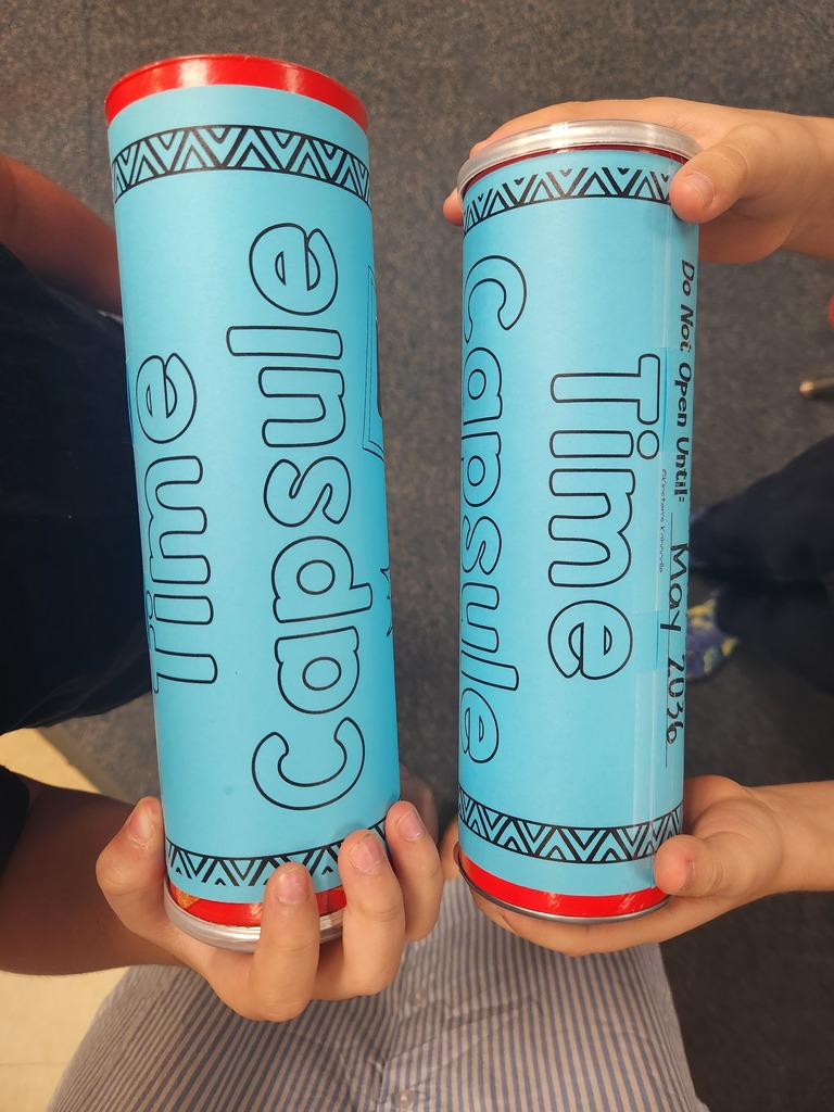 Two hands hold blue cylindrical objects with "Capsule Time" and decorative patterns on them. The background is a gray floor.