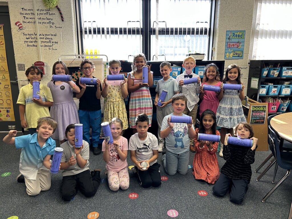 A classroom of children, seated and standing, holds purple tubes; some wear colorful dresses. A whiteboard with text is in the background.