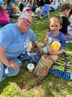 Woman and child sitting on grass, holding stuffed toy and cup. Background shows people in outdoor setting.