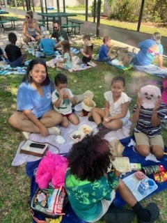 Adults and children are seated around picnic tables with food spread out. They are in a grassy area.