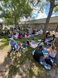 People sit on blankets on grass near trees and a building, with some in shaded areas.