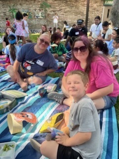 A family sitting on a picnic blanket, with a boy smiling, a man and a woman wearing sunglasses, surrounded by a group of people on a grassy area.