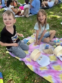 Two children sit on a blanket, smiling. A girl has a white shirt and a boy a black shirt. Various snacks surround them.