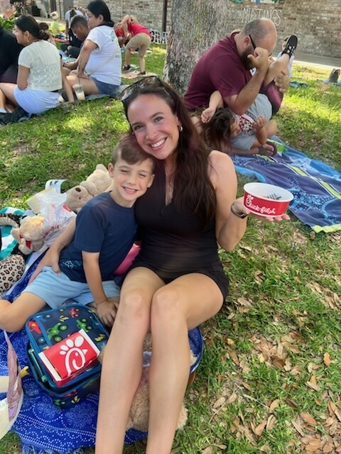 A woman and boy sit on a blanket with toys and food. Other families sit around them on grass.