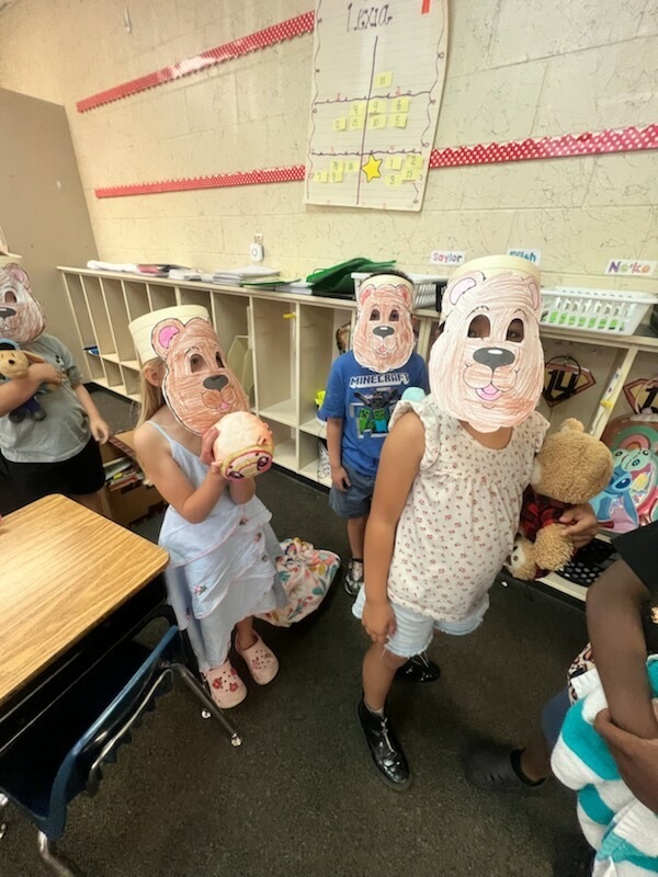Children wearing paper animal masks in a classroom with white shelves and a wooden desk.