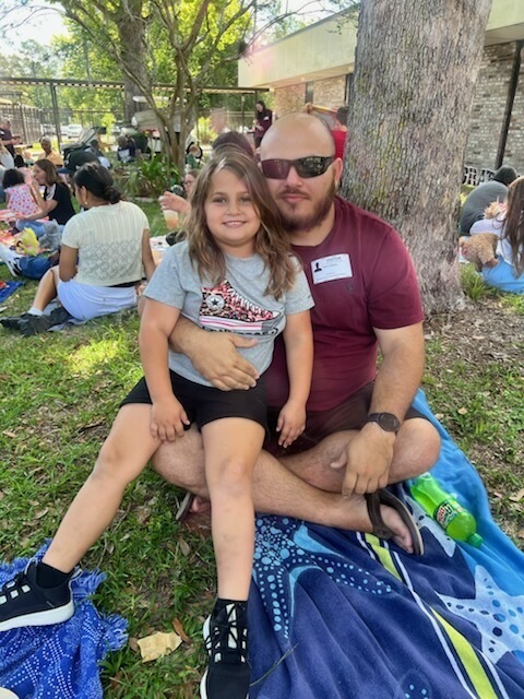 A man and a young girl are sitting on a blanket in a park. Behind them, a tree and a fence are visible.