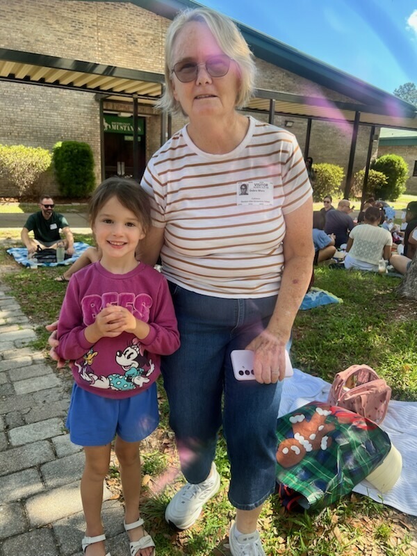 An older woman stands with a young girl in front of a building with people sitting on the grass.