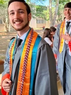 A man in a graduation gown stands smiling in front of trees and a building. Behind him, another person in a graduation gown walks.