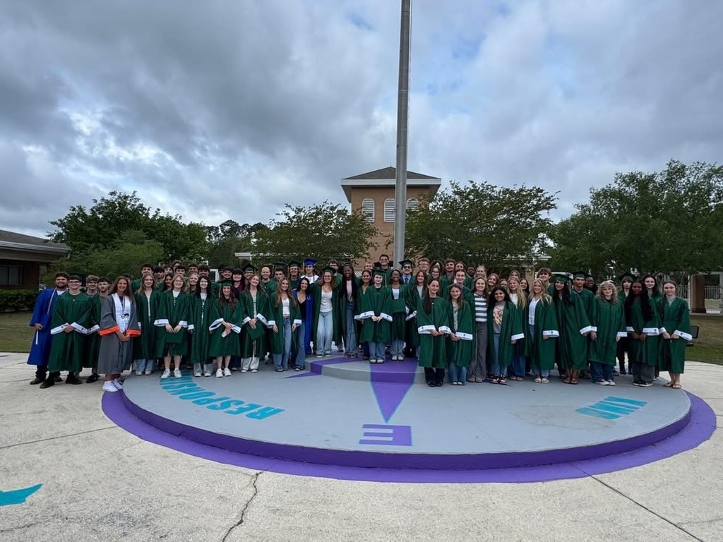 A large group of people in green gowns stand in a circle in front of a flagpole.