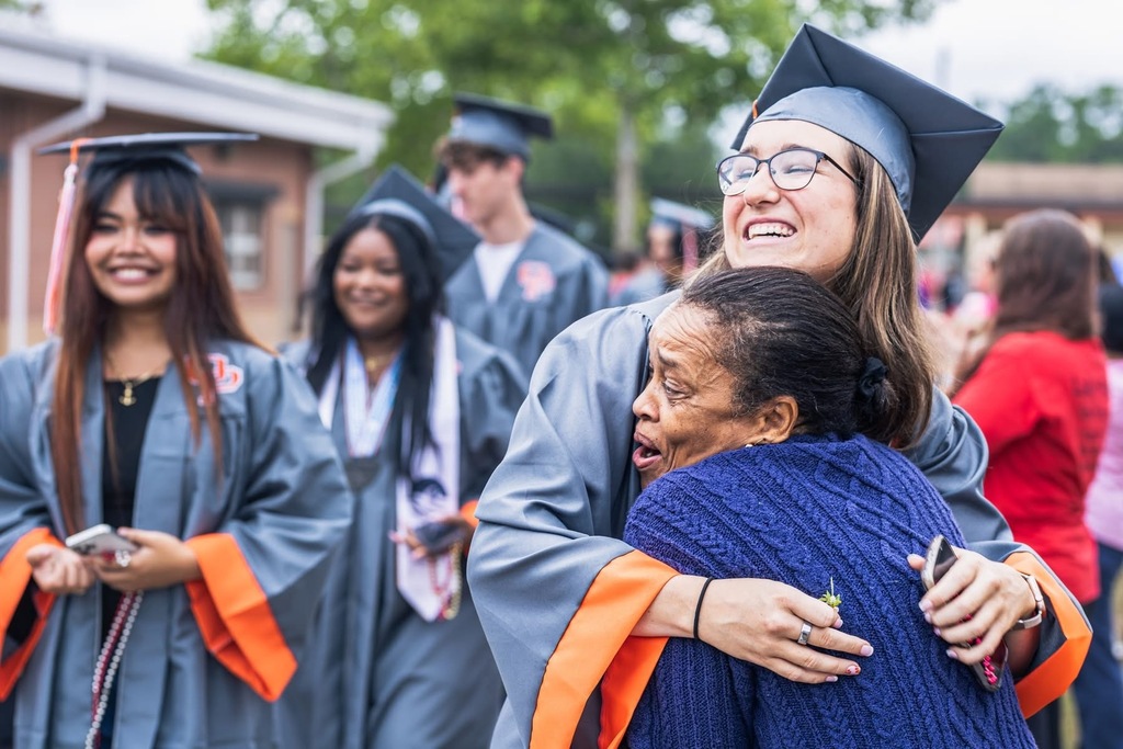 A group of women in graduation gowns, one hugging another while others look on at a school event.