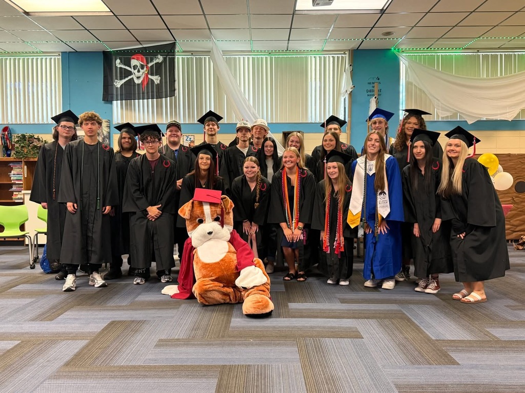 Group of people in graduation gowns and hats, one holding a mascot costume, standing in a decorated room.