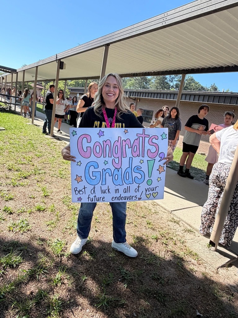 A woman holding a sign that reads "Congratulations Grads!" stands outside with people walking by.