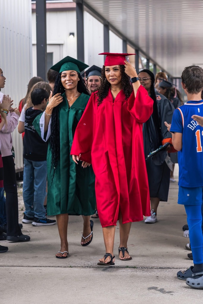 Two women in graduation gowns, one in red and the other in green, walk past others on a sidewalk.