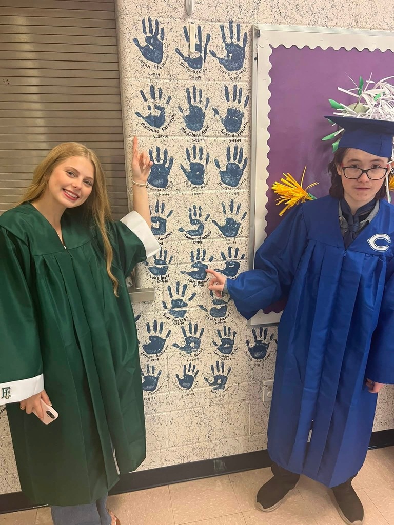 Two people in graduation gowns stand next to each other. A wall with handprints is behind them.