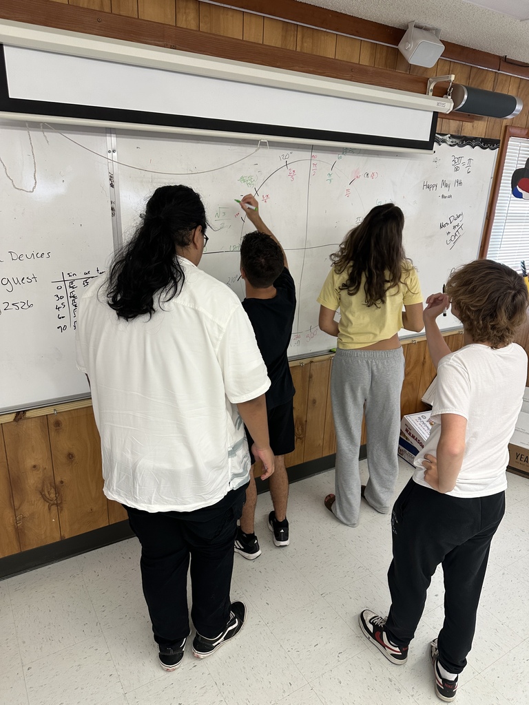 Four students are gathered around a whiteboard with various diagrams and text. Two are writing on the board while two are observing.