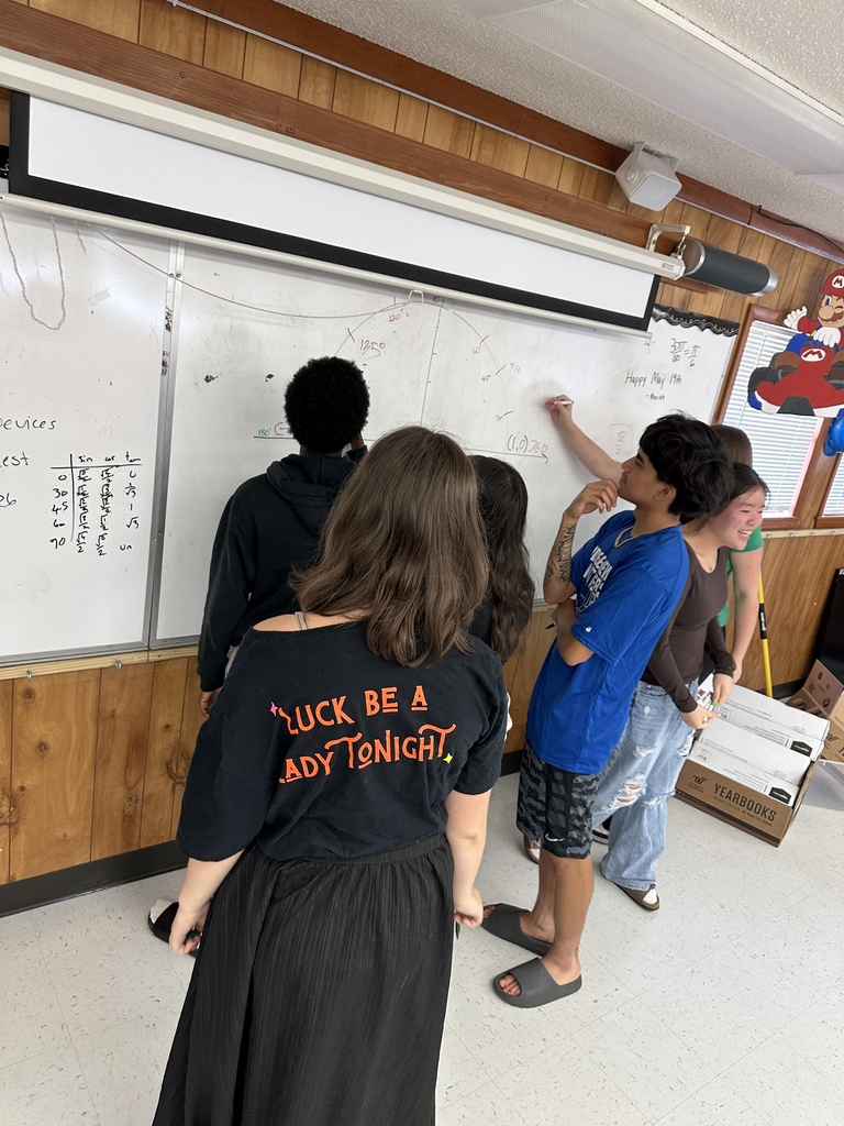 A group of students stands around a whiteboard. One person writes on the board while others watch.