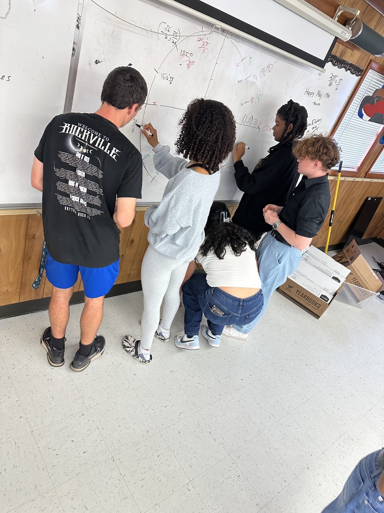 A group of students in a classroom are standing around a whiteboard, writing with markers.