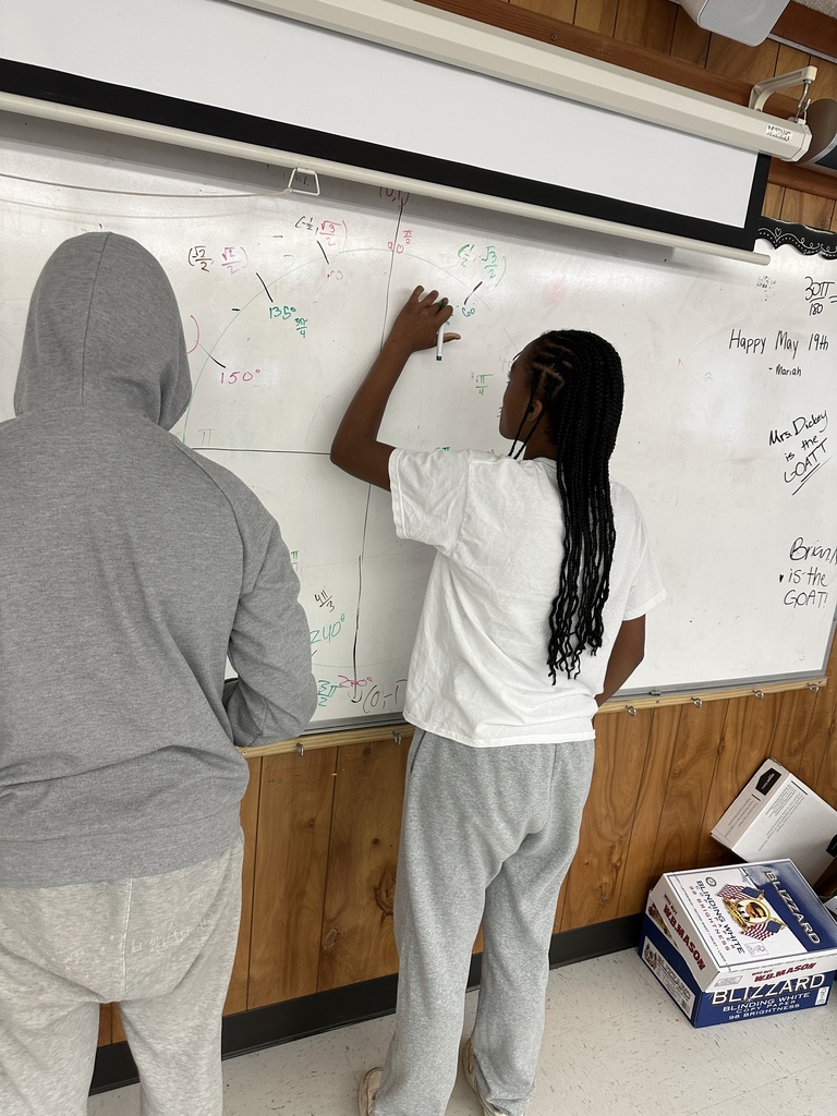 Two students, one with braided hair, write on a whiteboard. The room has wooden walls and a ceiling lamp.