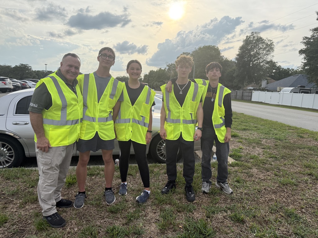 Five people in neon yellow vests and sneakers stand in grass, with a white car behind them.