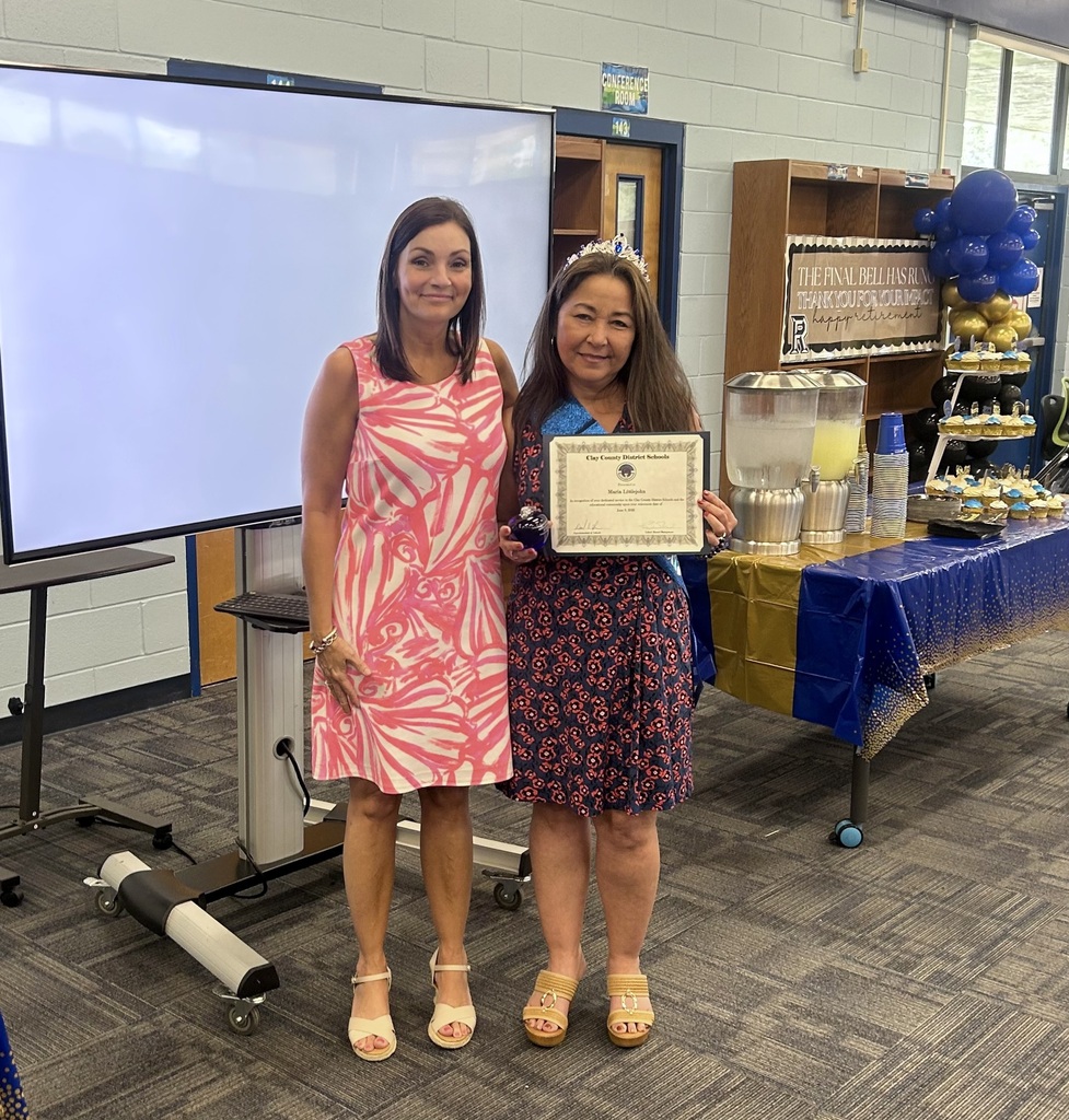 Two women stand in a room, one holding a framed certificate. Behind them, a projector screen is on a stand, and a table holds a blue tablecloth.