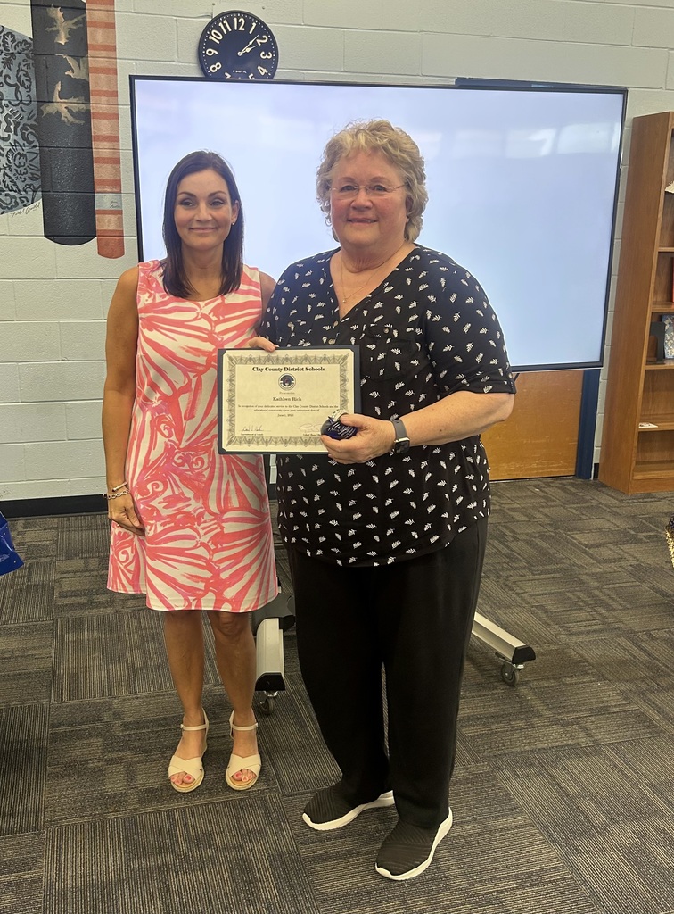 Two women in a room with a clock and a projector screen. One woman holds an award certificate.
