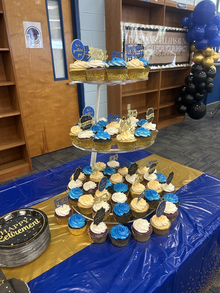 A table with a blue tablecloth displays a three-tiered stand holding cupcakes. The cupcakes are frosted blue and gold.