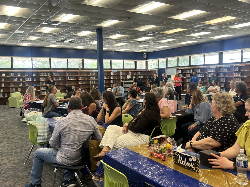 People in a library, sitting at tables with refreshments, some standing and talking near shelves with books.