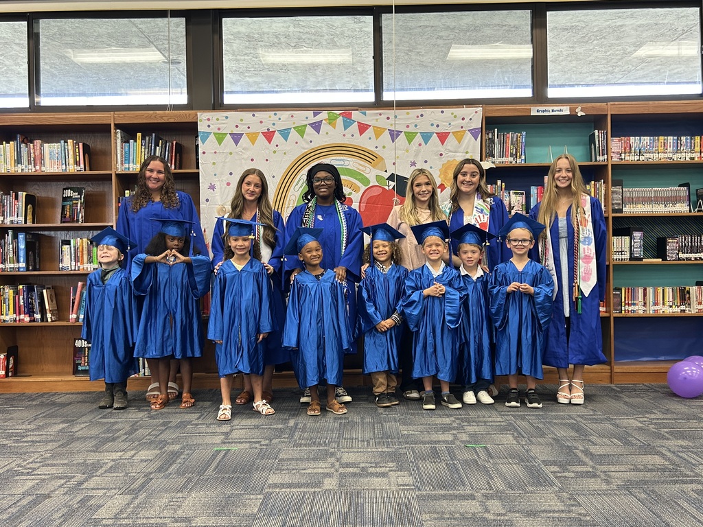 Group of children and adults in blue graduation gowns standing in a library with bookshelves and balloons.
