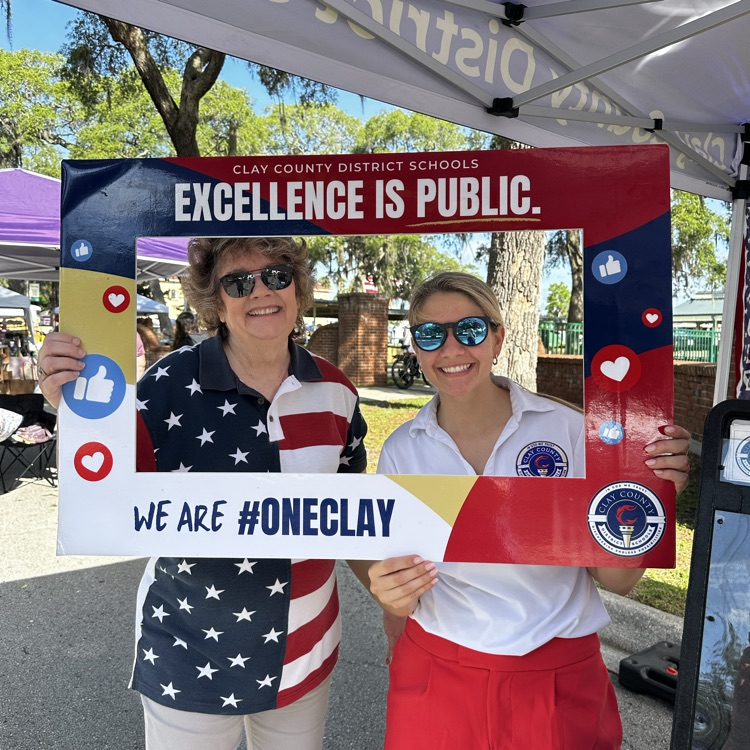 Two women holding a sign that reads “excellence is public” in front of a park.