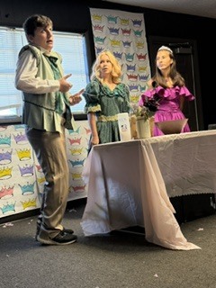 Three people, one male and two females, stand in front of a table. Behind them is a colorful banner.