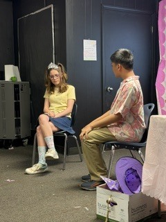 Two people seated in chairs, one with glasses and a yellow shirt, the other in a patterned shirt. A purple hat rests on a box.