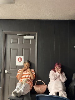 Two women sit near a door. One wears orange and white, the other pink. A pink basket rests between them.