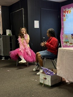 Two girls sit on chairs. One wears a pink dress and scarf and holds a microphone. The other wears a red jacket. They face each other, with a box on the floor.
