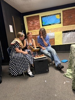 Three people in casual attire sit on a bench. Two of them converse, while the third holds a book. They are in a room with a yellow wall.