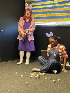 A woman in bunny ears stands next to a girl in overalls who is sitting on the floor with dice.