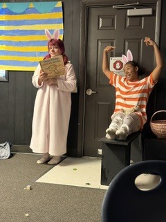 Two people in bunny costumes standing in a room. One person holds a book, and another raises arms near a door.