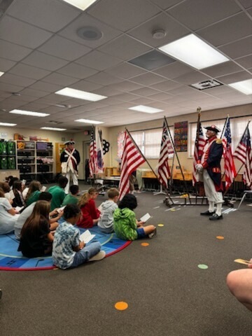 Classroom with children seated in a circle. Two adults in historical costumes stand in the center with flags.
