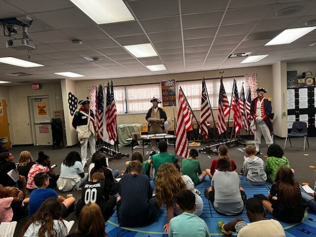 People dressed in colonial attire stand on stage with flags behind them, engaging with a seated audience in a classroom.