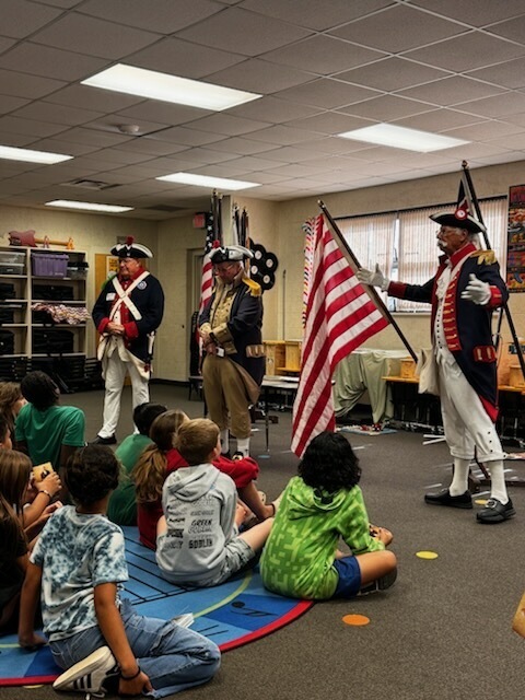 Several children sit on the floor while adults in Revolutionary War-era costumes hold flags and talk.