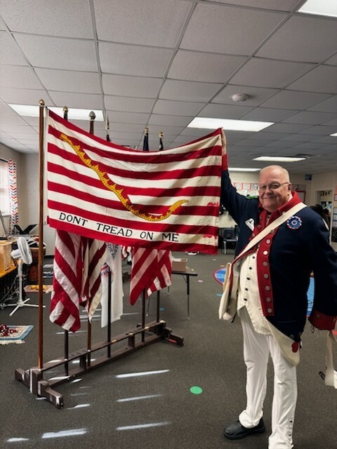 A man in historical military uniform holding a flag with text. Several flags and furniture in the background.