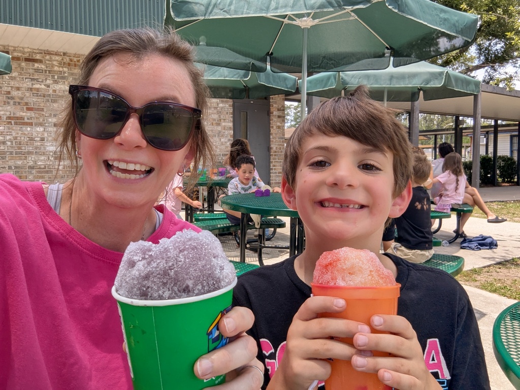 A woman and a boy hold green and orange shaved ice cups, smiling at the camera. Behind them, tables with umbrellas, children sitting, and a brick building.