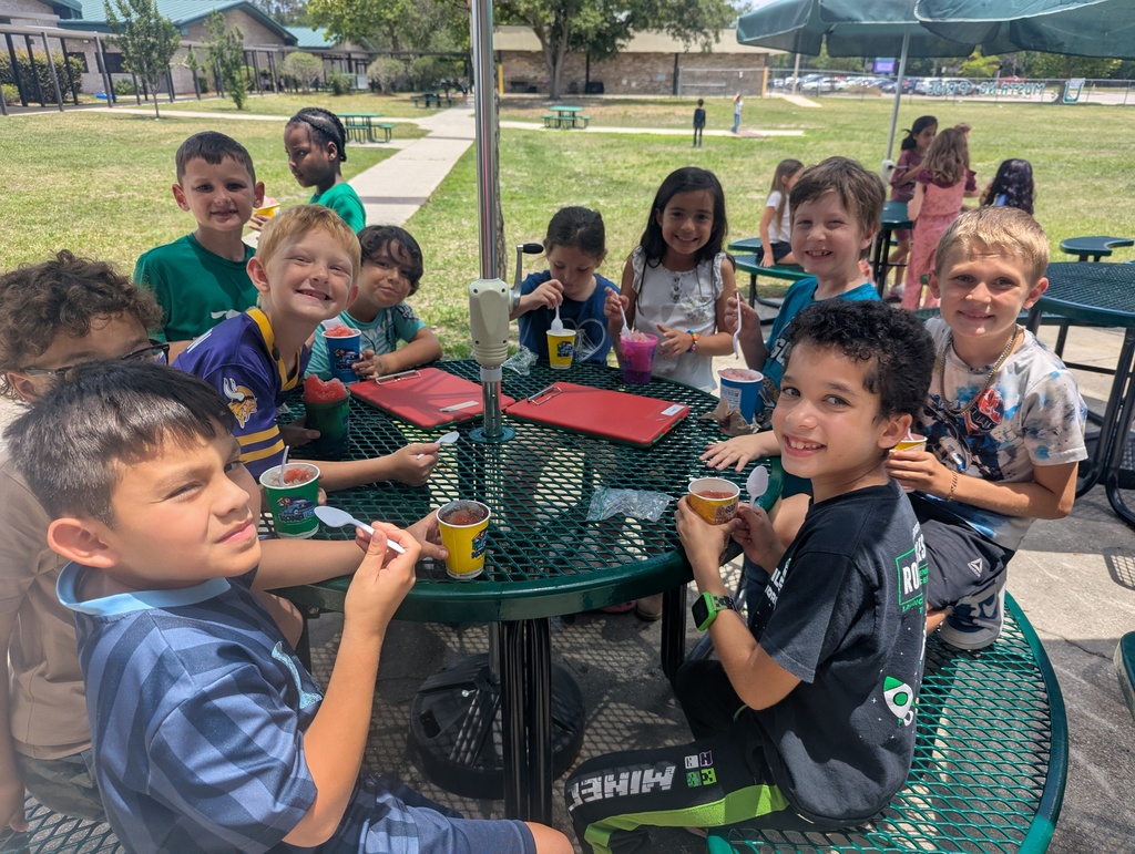 Kids sitting at a table eating snacks. Umbrellas provide shade. A grassy field is in the background.