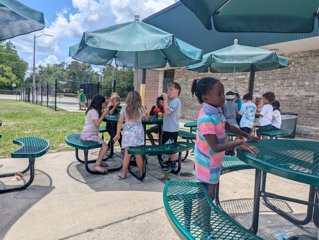 Several children sit at picnic tables under umbrellas in a shaded outdoor area with grass.
