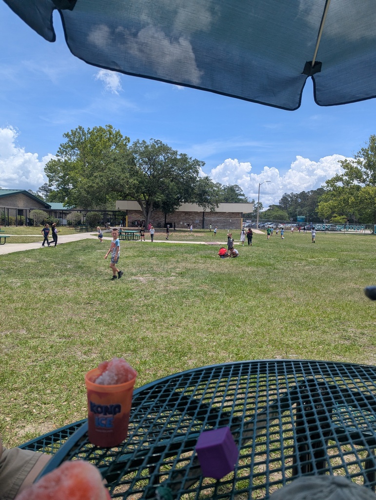 A park with people in the distance. A table with a drink container and a small cube.