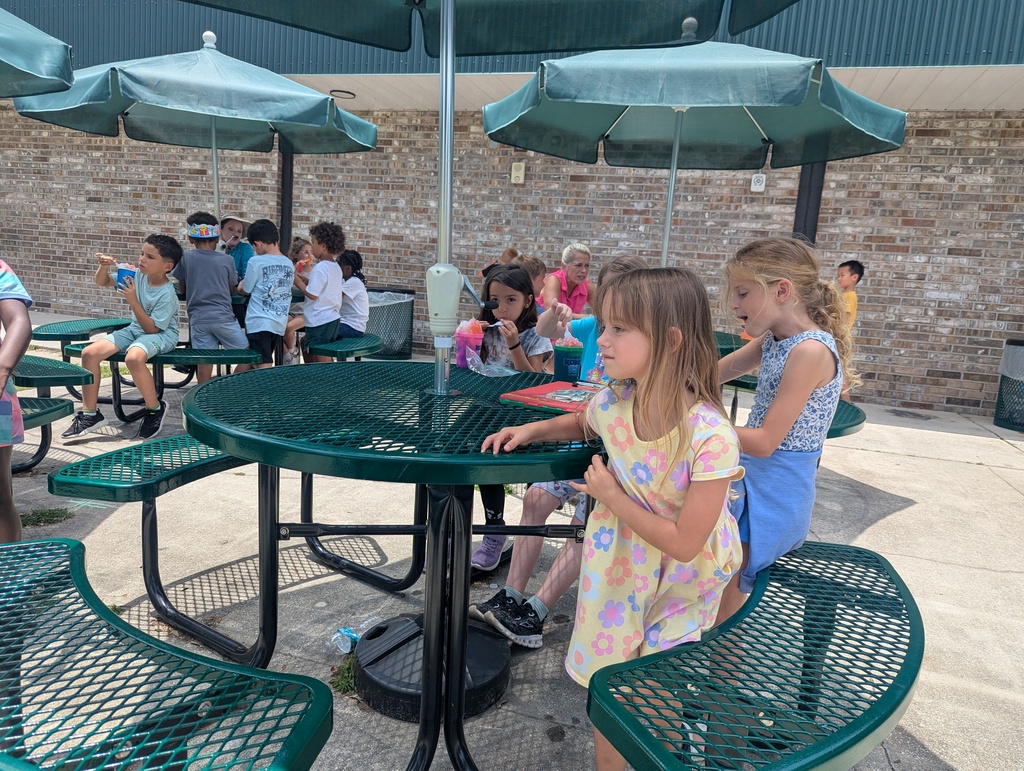 A group of children in a picnic area. Some are seated, while others stand around a table with green benches and green umbrellas.
