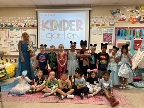Group photo of young children dressed in costumes in a classroom with a projector screen showing "Kindergarten."