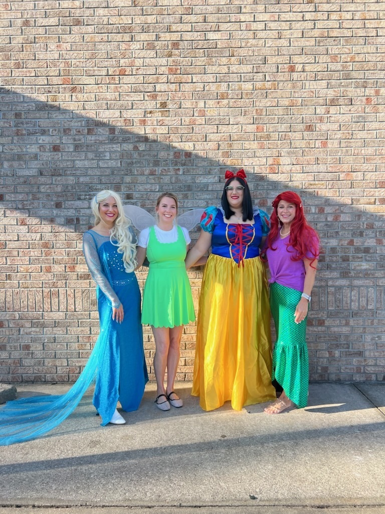 Four women dressed in princess costumes stand next to a brick wall, posing for a picture.
