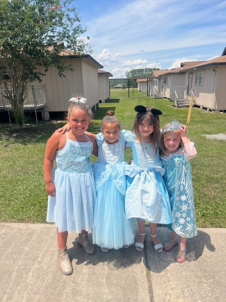 Four young girls in blue dresses and sandals, two with mouse ears, standing in front of houses.