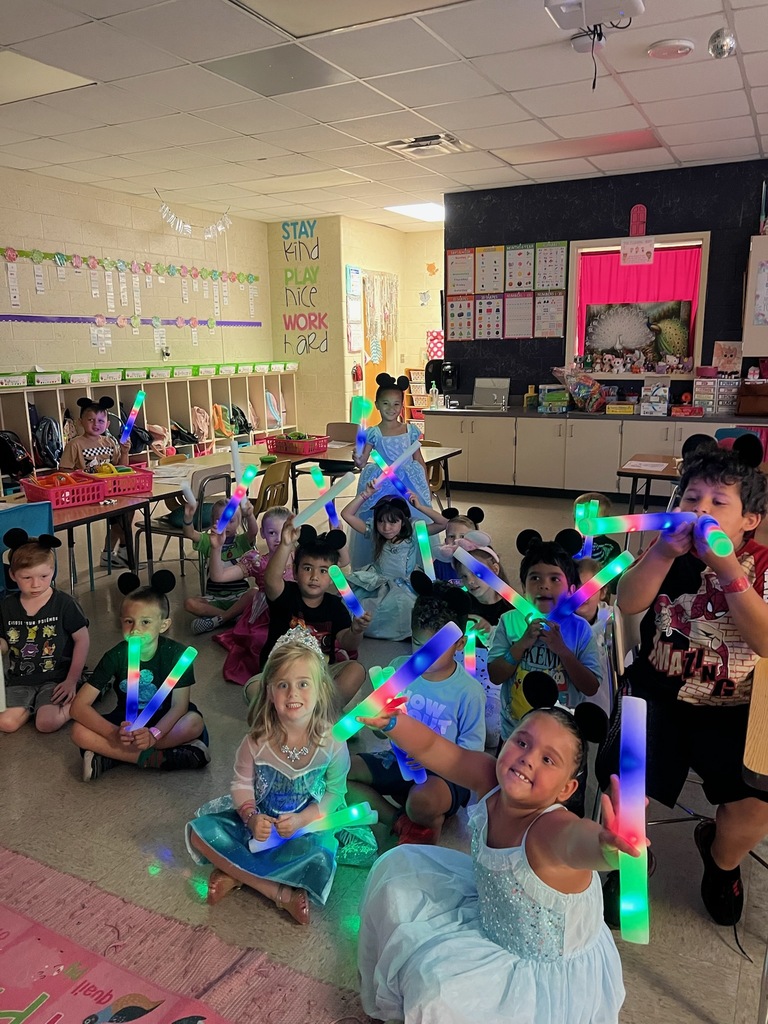 Kids wearing costumes and holding light-up wands sit on a pink rug in a classroom.